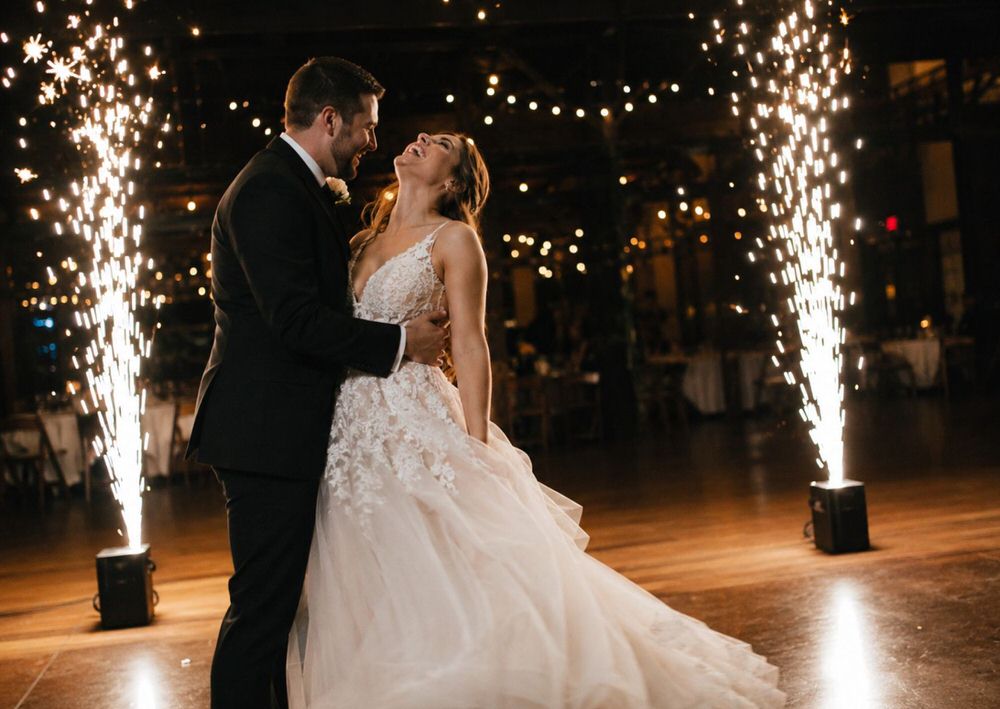 Bride and groom laughing during their first dance, flanked by cold spark fountains at an indoor wedding reception