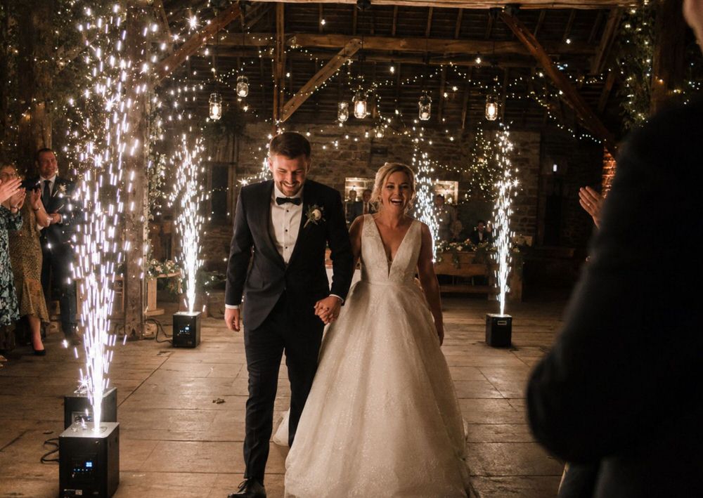Newlyweds walking hand in hand through a cold spark entrance corridor as guests applaud at a barn wedding venue